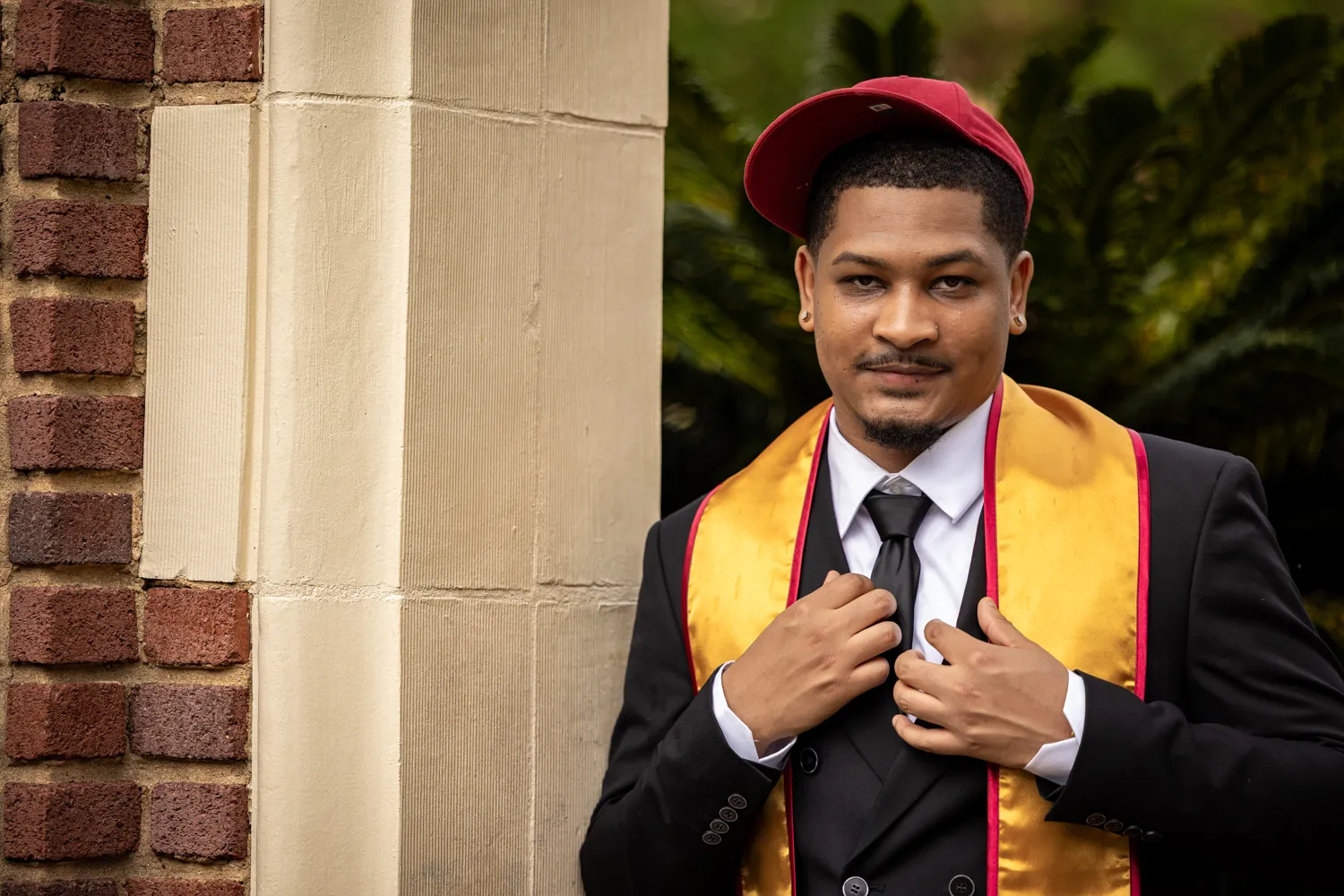 A close-up portrait of a graduate wearing a backward garnet cap, adjusting the lapels of his black suit jacket. His gold FSU graduation stole is prominently displayed, and a brick pillar is visible on the left.
